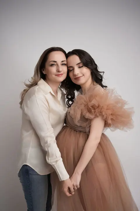 Две жени буза до буза, хванати за ръце; бременната е в прасковена тюлена рокля