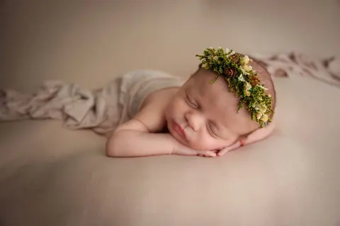 Новородено с венче от зеленина и жълти цветчета спи с глава върху ръчичките