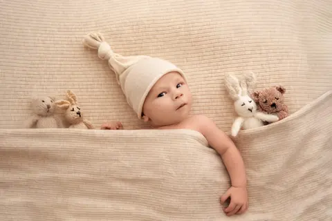 Будно бебе в кремава шапчица с възел наднича изпод завивката до плетени зайчета