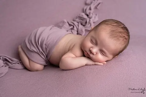 Новородено спи на коремче с ръчичка под бузката, покрито с люляков плат